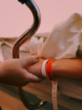 A child in hospital. Their hand is being held by an older person. The child's wrist has lots of bands on it from previous surgeries and hospital stays. The background can barely be seen, but it is a light pink.