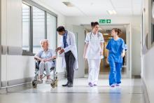 Four people in a hospital corridor. A doctor in a white coat speaking to an elderly woman in a wheelchair, and two nurses.