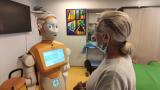 An orange robot called Ari speaks to a white woman.