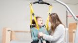 A young girl with a disability being lifted with a harness. Her carer guides her from the bed, looking at her and smiling.