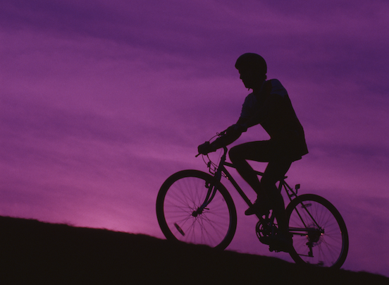A silhouette of a person cycling up a hill against a purple sky