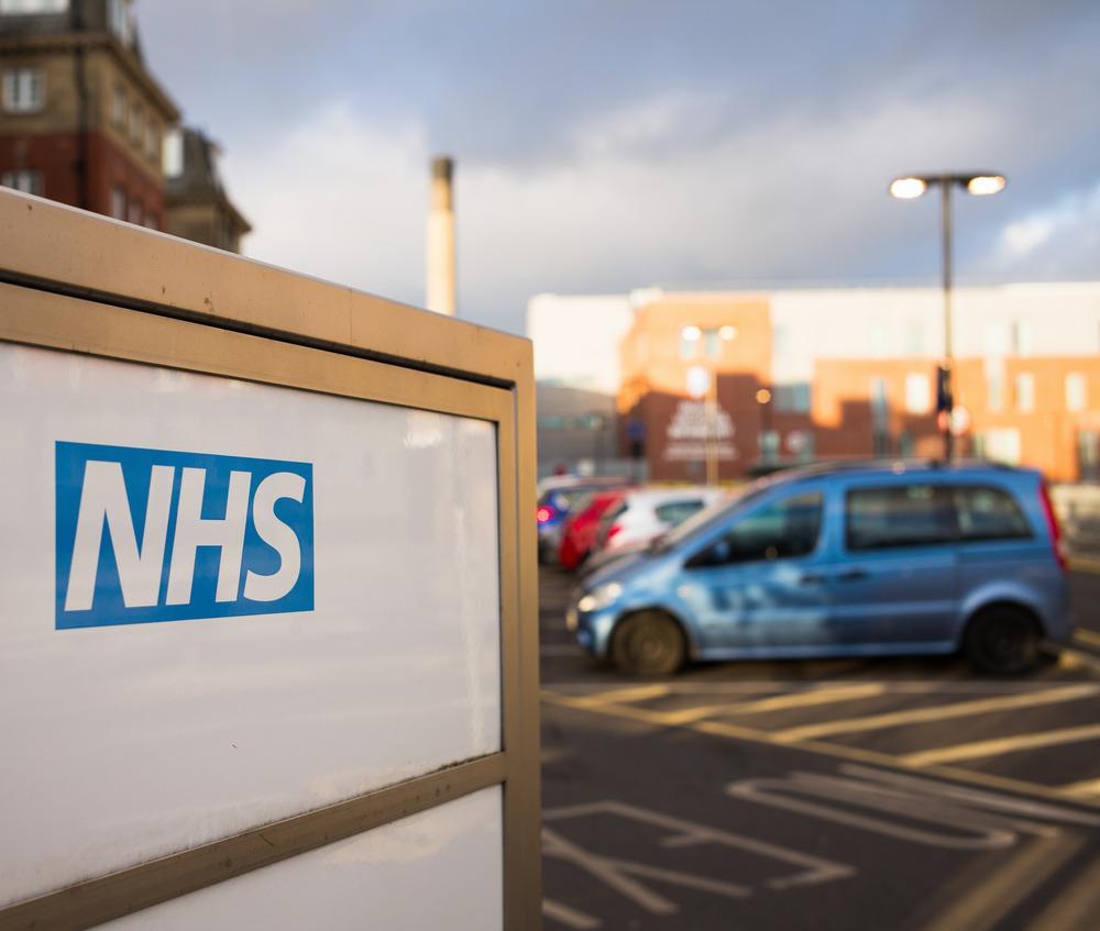 A sign says 'NHS'. A car park in the background is out of focus with a blue car visible.