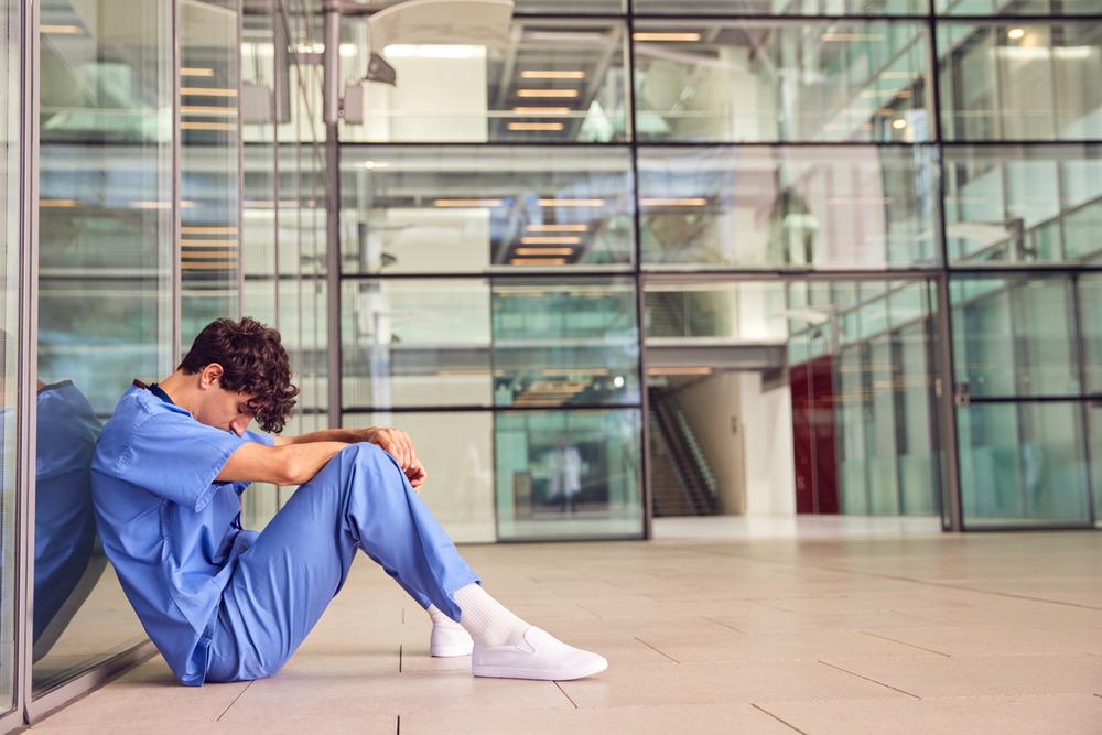 A junior doctor leans against a modern hospital building.