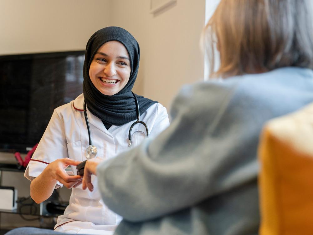 A young doctor talks to a patient, smiling. She has a stethoscope around her neck.